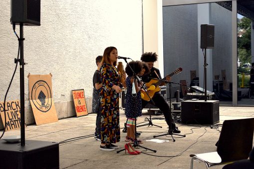 Ein Schwarzer Gitarrist und eine Weiße Sängerin musizieren zusammen mit zwei Schwarzen Kindern auf einer Bühne.